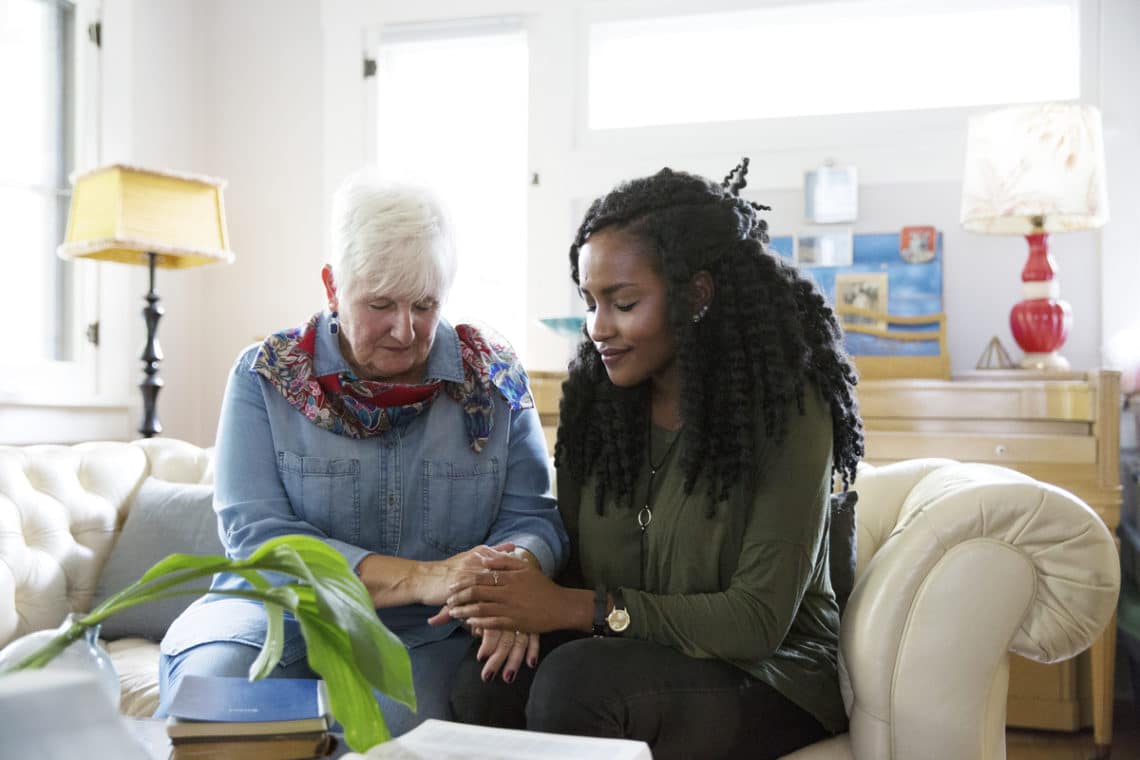 A mature woman and young woman sit together on a sofa and pray holding hands